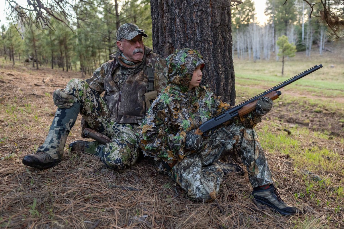 A man and teenager dressed in camouflage clothes sit beneath a large pine tree in the morning, the teenager is holding a shotgun.