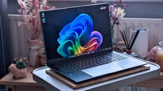 An Acer Swift Go 16 AI laptop sits on a small table, displaying the Windows 11 desktop. The keyboard and touchpad are visible, and the background includes decorative items like dried flowers.