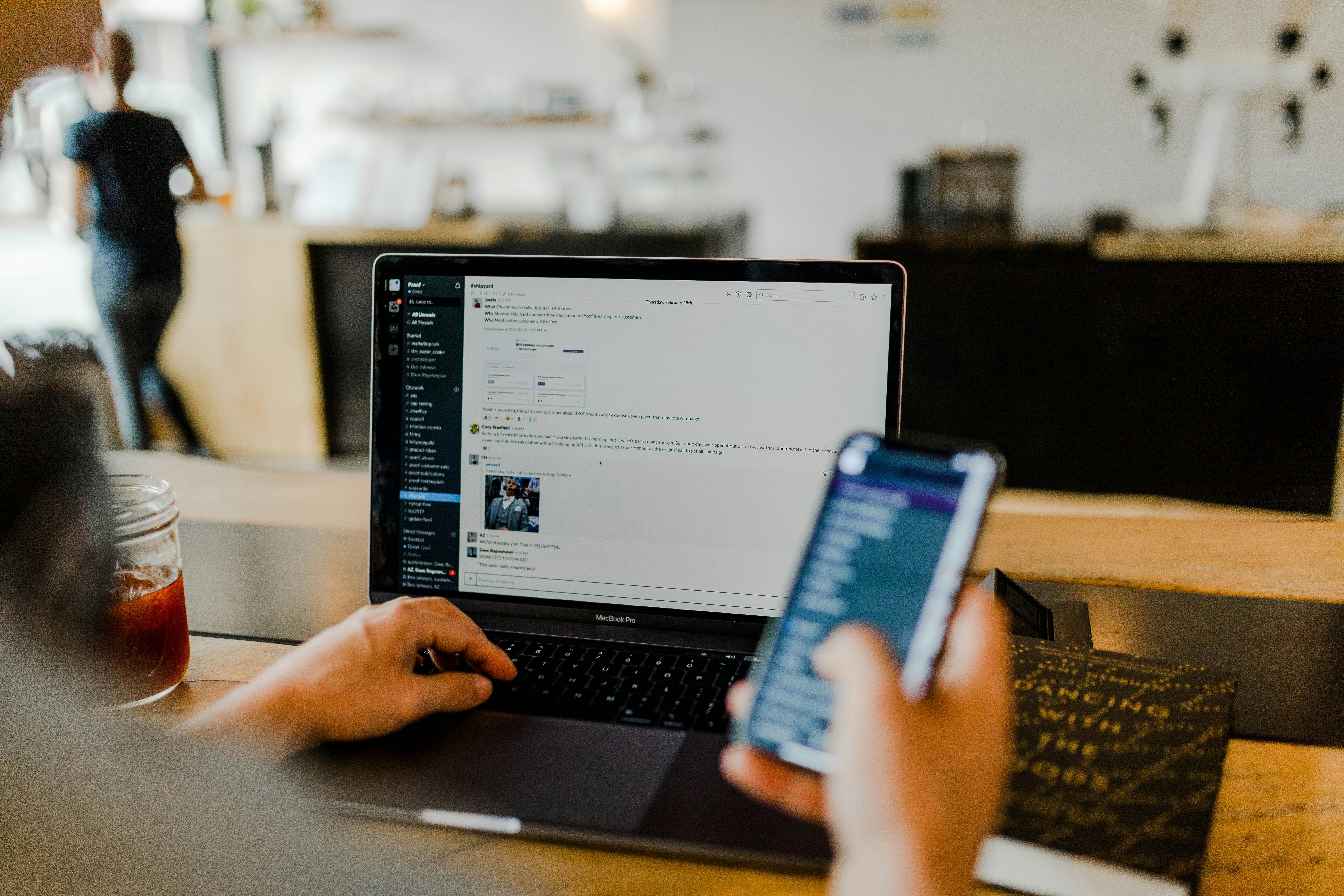 a stock photo of someone looking at their phone while working on their computer with Slack open.