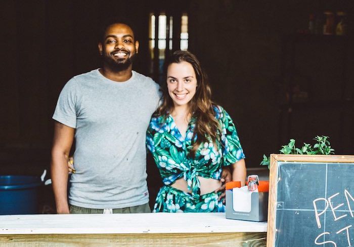“Ephream and Jocey beam behind a bar counter, welcoming guests. In front of of the bar counter leans a chalkboard sign with the venue's name and a list of drinks.