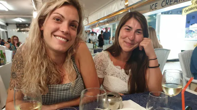 Céline, de cabelos longos e loiros, sorri para a câmera usando uma blusa de alcinha xadrez. Justine, de cabelos longos castanho-escuros, sorri com uma das mãos apoiada no rosto. Ela veste uma blusa branca com acabamento em renda