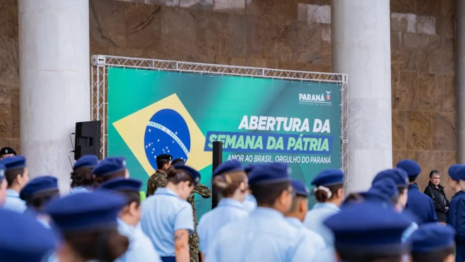 Estudantes em escola cívico-militar no Paraná, com placa ao fundo que contém bandeira do Brasil e dizeres "Abertura da Semana da Pátria - Amor ao Brasil, orgulho do Paraná"