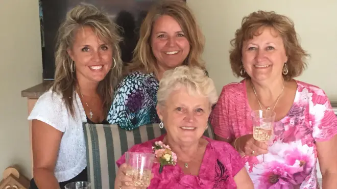 Anita LeBrun, de 82 años, con una blusa de flores rosa intenso, sonríe mientras sostiene una copa de vino blanco, rodeada por las mujeres de su familia.