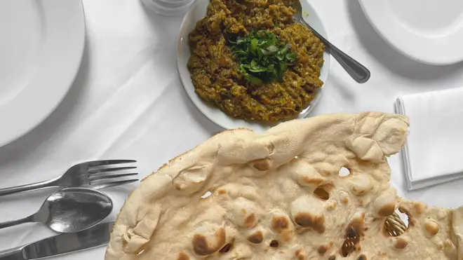 نان تافتون و کشک بادمجان روی یک رومیزی سفید در کنار قاشق، چنگال و کارد و چند بشقاب خالی.