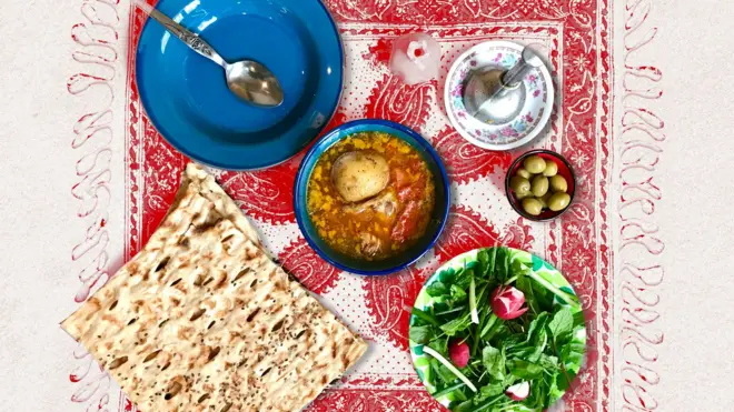 نان سنگک و آبگوشت، سبزی خوردن و زیتون روی یک رومیزی قرمز در کنار گوشت کوب و ظرف خالی و یک قاشق.