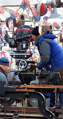 大型の映画用カメラを覗き込む、青いジャケットと黒い帽子の男性。頭上には赤白青の幔幕が飾られている。