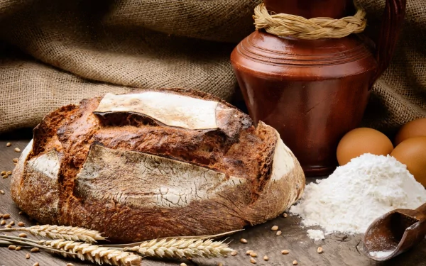HD PC desktop wallpaper showing a rustic loaf of bread surrounded by flour, eggs, wheat stalks, and a clay jug on a burlap surface.