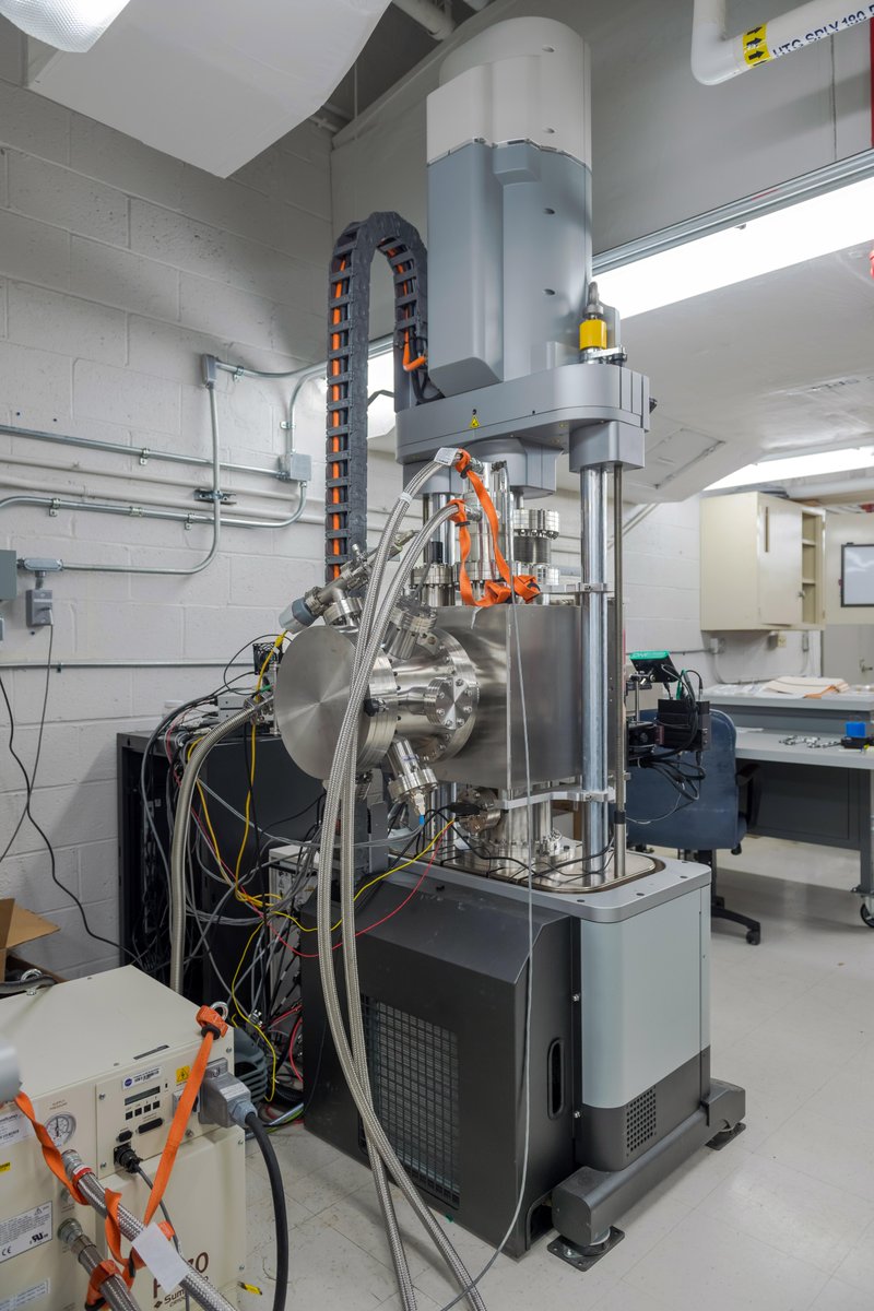 A tall, industrial machine with metallic and gray components stands in a laboratory. Multiple cables and tubes connect to the central chamber, which is cylindrical. Some cables are silver and braided, while others are orange or yellow. The test rig has vertical support columns and a black base. In the background there are white brick walls, ceiling ducts, and a work station with a desk and chair. Credit: NASA/Steven Logan